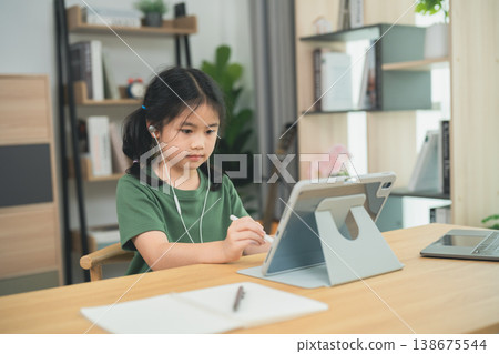 Young girl engaged in online learning at home, focusing on digital education, using tablet, wearing headphones, seated at wooden desk in modern interior 138675544