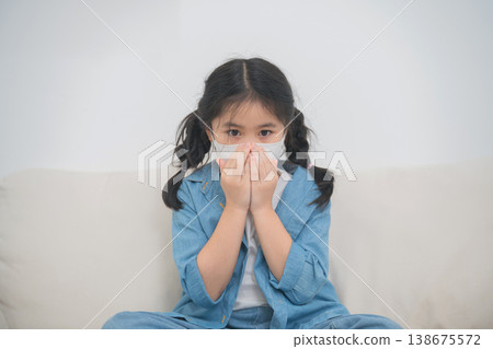 Young girl wearing a protective mask sitting on a couch at home, demonstrating caution and concern during health crisis or pandemic situation 138675572