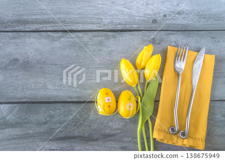 Easter dinner concept; place setting with forged fork and knife, bouquet of yellow tulps and easter eggs on a gray wooden background; copy space 138675949