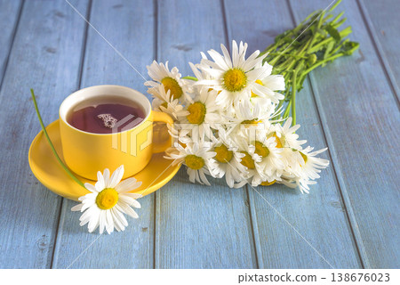Yellow cup of tea and bouquet of white daisies flowers on blue wooden background; close up Yellow cup of tea and bouquet of white daisies flowers on blue wooden background; close up 138676023