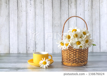 Bouquet of white daisies or chamomile flowers in wicker basket and yellow cup of tea on a wooden background; copy space Bouquet of white daisies or chamomile flowers in wicker basket and yellow cup of tea on a wooden background; copy space 138676024