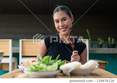 Young Thai Woman Holding Grilled Liver Skewer with Traditional Isan Thai Food at Restaurant Table 138676116