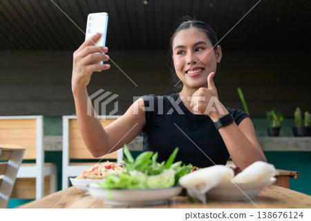 Happy Thai Woman Giving Thumbs Up Taking Selfie with Smartphone at Restaurant with Thai Food Happy Thai Woman Giving Thumbs Up Taking Selfie with Smartphone at Restaurant with Thai Food 138676124