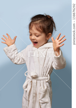 little girl in a white bathrobe looking surprised and excited with hands raised on blue background 138676292
