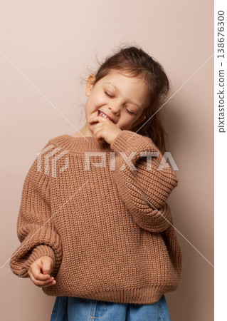 cute little child girl smiling, biting finger, expressing joy, wearing brown knit sweater on beige background cute little child girl smiling, biting finger, expressing joy, wearing brown knit sweater on beige background 138676300