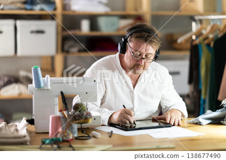 Middle-aged male dressmaker wearing earphones drawing on tablet in sewing workshop Middle-aged male dressmaker wearing earphones drawing on tablet in sewing workshop 138677490