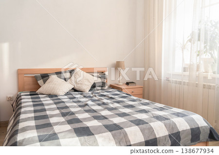 Cozy bedroom interior featuring a bed with plaid blanket, soft accent pillows, and bright sunlight streaming through a window 138677934