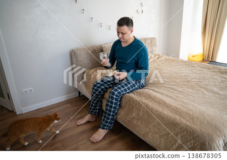 A 35 year old caucasian man sits on his bed in his bedroom with pills and water in his hands A 35 year old caucasian man sits on his bed in his bedroom with pills and water in his hands 138678305