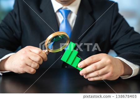 Businessman analyzes a falling chart with a magnifying glass. Examining financial decline Businessman analyzes a falling chart with a magnifying glass. Examining financial decline 138678403