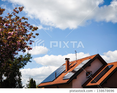 Solar panels installed on a modern house roof surrounded by blooming trees under a bright blue sky with fluffy clouds showcasing renewable energy concept 138678696