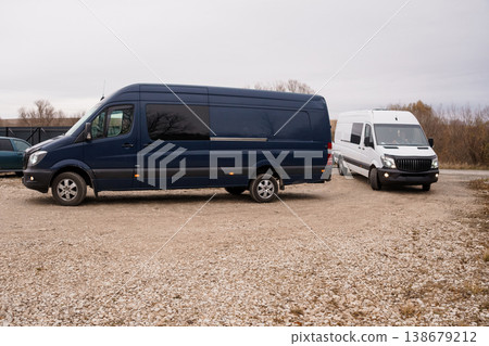 Dark Blue Large Cargo Van and White Transport Van Parked on Gravel Lot Under Overcast Sky. Dark Blue Large Cargo Van and White Transport Van Parked on Gravel Lot Under Overcast Sky. 138679212