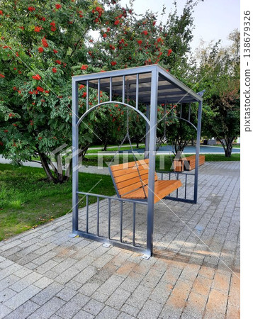 Modern wooden park swing bench on a metal frame surrounded by green trees in a city garden. Modern wooden park swing bench on a metal frame surrounded by green trees in a city garden. 138679326