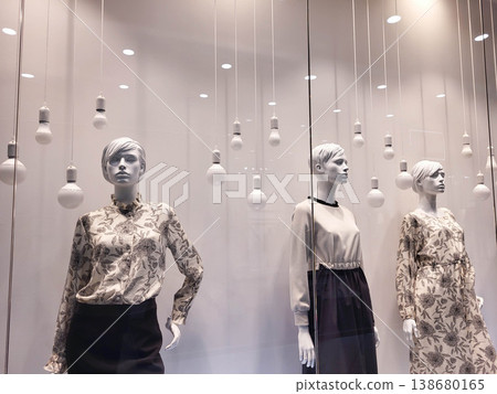 In the store window, there are three female mannequins dressed in clothes and hanging white light bulbs. 138680165