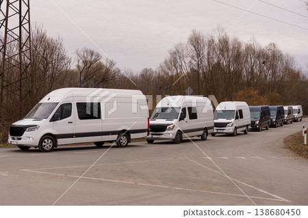 Fleet of white delivery vans parked in a row on an asphalt road with autumn trees in background. 138680450