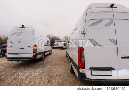 Two white commercial transport vans parked on a gravel lot under a cloudy overcast sky. Two white commercial transport vans parked on a gravel lot under a cloudy overcast sky. 138680461