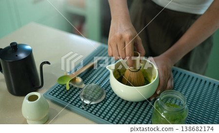 Hands whisking matcha powder in ceramic bowl on kitchen counter. 138682174