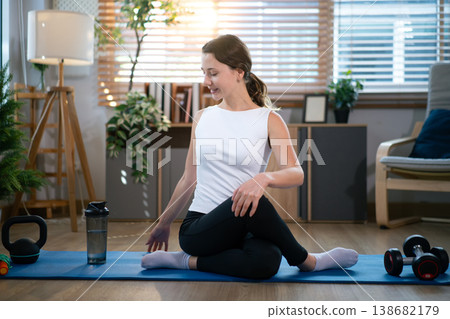 Smiling woman sitting on yoga mat after indoor exercise session 138682179