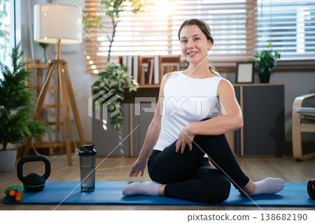 Smiling woman sitting on yoga mat after indoor exercise session Smiling woman sitting on yoga mat after indoor exercise session 138682190