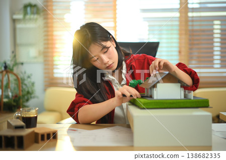 Female architect examining house model at office desk, Professional architect reviewing architectural house model Female architect examining house model at office desk, Professional architect reviewing architectural house model 138682335