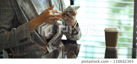 Young businesswoman in suit sitting at table with paper cup of coffee and using mobile phone Young businesswoman in suit sitting at table with paper cup of coffee and using mobile phone 138682365