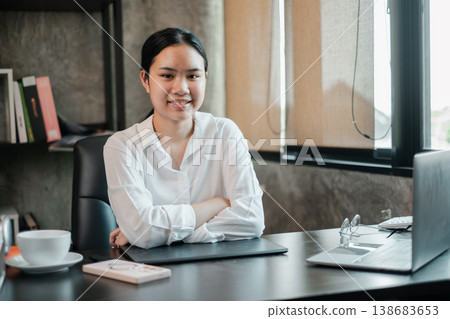 Young woman in a white shirt sitting at a desk in a modern office, smiling confidently with a laptop and documents. 138683653