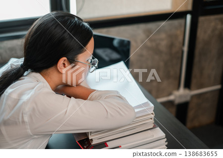 Young woman in glasses leans on books, reflecting in a modern office. Ideal for themes of education, work, or contemplation. 138683655