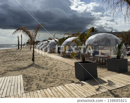 Beachside dining domes on sandy shore with palm trees under partly cloudy sky background 138685821