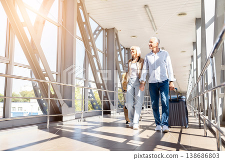 Mature couple walking together in airport terminal with luggage, smiling and relaxed Mature couple walking together in airport terminal with luggage, smiling and relaxed 138686803