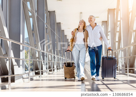 Mature couple walking together through sunlit airport terminal, pulling suitcases Mature couple walking together through sunlit airport terminal, pulling suitcases 138686854