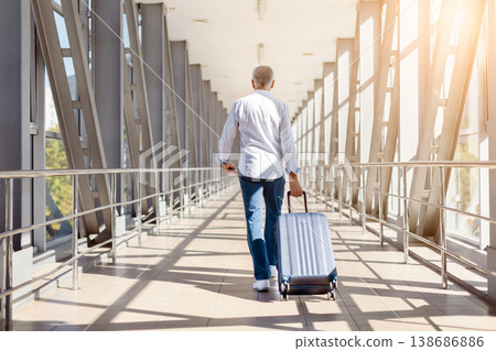 Mature man walking away in airport terminal pulling suitcase, rear view Mature man walking away in airport terminal pulling suitcase, rear view 138686886