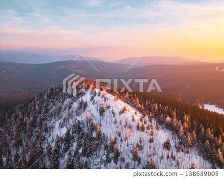 Bukovec Mountain in the Jizera Mountain range. The drone captures the landscape of trees and snow with a warm light from the setting sun, showcasing nature's features. Bukovec Mountain in the Jizera Mountain range. The drone captures the landscape of trees and snow with a warm light from the setting sun, showcasing nature's features. 138689005