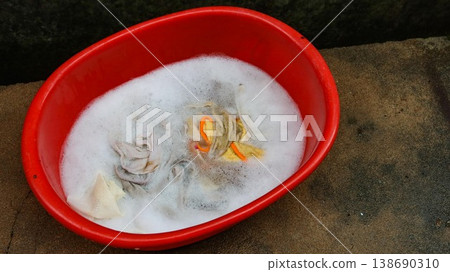 close up of a red plastic basin filled with soapy water and submerged fabrics for hand washing clothes on the ground with copyspace 138690310