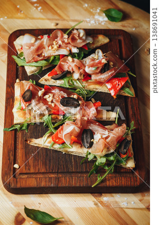 Italian flatbread appetizers with prosciutto ham, fresh arugula, black olives and pine nuts served on a dark wooden board. Italian flatbread appetizers with prosciutto ham, fresh arugula, black olives and pine nuts served on a dark wooden board. 138691041