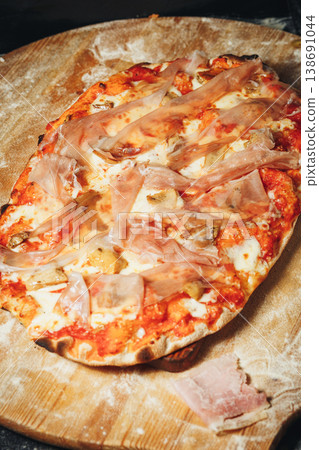 Traditional Italian pinsa romana topped with tomato sauce melted mozzarella savory mushrooms and thin slices of prosciutto ham on a floured wooden board. Traditional Italian pinsa romana topped with tomato sauce melted mozzarella savory mushrooms and thin slices of prosciutto ham on a floured wooden board. 138691044
