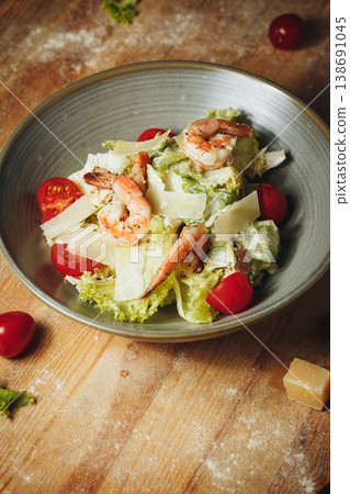 Fresh Caesar salad with grilled shrimps, cherry tomatoes, parmesan shavings and crisp lettuce in a ceramic bowl on a rustic wooden table. 138691045