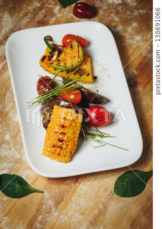 Gourmet grilled vegetables platter featuring sweet corn zucchini bell peppers and cherry tomatoes served on a white plate over a rustic wooden background. Gourmet grilled vegetables platter featuring sweet corn zucchini bell peppers and cherry tomatoes served on a white plate over a rustic wooden background. 138691066