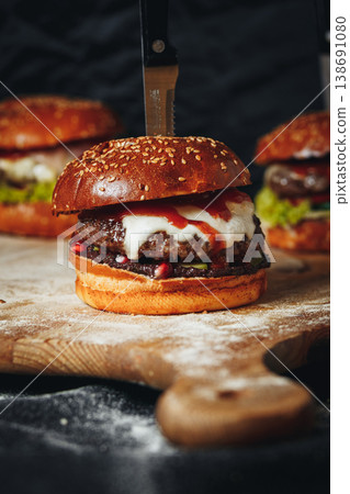 Gourmet beef burger with melted cheese and pomegranate seeds served on a rustic wooden board with a knife stuck through the bun. Gourmet beef burger with melted cheese and pomegranate seeds served on a rustic wooden board with a knife stuck through the bun. 138691080