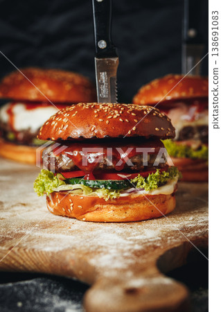 Gourmet beef burger with melted cheese red sauce and fresh vegetables served on a rustic wooden board with a knife stuck through the bun. 138691083