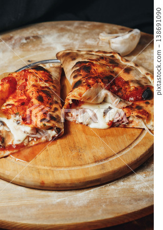 Traditional Italian calzone pizza cut in half with melted mozzarella cheese ham and mushrooms on a rustic wooden board with flour. 138691090