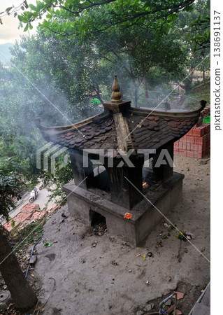 An outdoor incense burner with smoke rising, set against lush greenery and a street scene with motorbikes in Vietnam An outdoor incense burner with smoke rising, set against lush greenery and a street scene with motorbikes in Vietnam 138691137