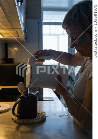 An elderly woman with dark circles under her eyes pours hot water from an electric kettle into teapot in her kitchen, enjoying her morning routine. Real people, life of an elderly person An elderly woman with dark circles under her eyes pours hot water from an electric kettle into teapot in her kitchen, enjoying her morning routine. Real people, life of an elderly person 138691979