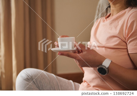 Close-up female hands holding a white ceramic cup of hot coffee drink on a saucer Close-up female hands holding a white ceramic cup of hot coffee drink on a saucer 138692724