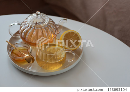 Glass teapot with amber tea, teacup with lemon slice, and bowl of honey with golden spoon on white table in cozy living room Glass teapot with amber tea, teacup with lemon slice, and bowl of honey with golden spoon on white table in cozy living room 138693104