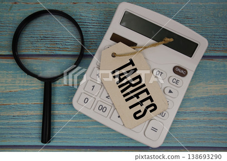 Price tag with "TARIFFS" on calculator and magnifying glass on a rustic blue wooden surface Price tag with "TARIFFS" on calculator and magnifying glass on a rustic blue wooden surface 138693290