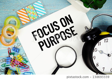 Focus On Purpose text on a paper surrounded by office supplies, a magnifying glass, and a clock on a blue wooden surface Focus On Purpose text on a paper surrounded by office supplies, a magnifying glass, and a clock on a blue wooden surface 138693339