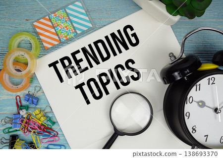 Trending Topics text on a paper surrounded by office supplies, a magnifying glass, and a clock on a blue wooden surface Trending Topics text on a paper surrounded by office supplies, a magnifying glass, and a clock on a blue wooden surface 138693370