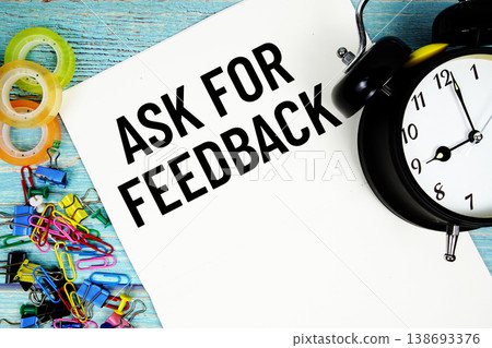 Ask For Feedback text on a paper surrounded by office supplies and a clock on a blue wooden surface 138693376