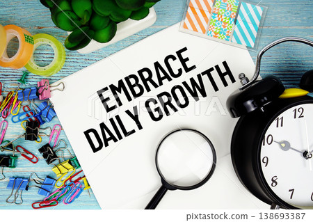 Embrace Daily Growth text on a paper surrounded by office supplies, a magnifying glass, and a clock on a blue wooden surface 138693387