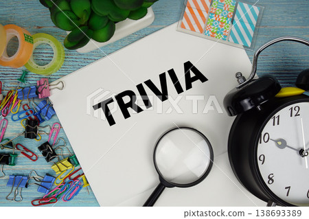 Trivia text on a paper surrounded by office supplies, a magnifying glass, and a clock on a blue wooden surface 138693389