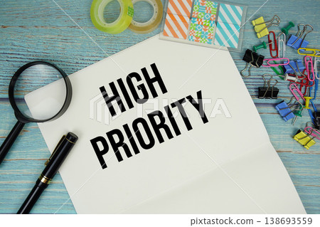 High Priority text on a paper surrounded by office supplies and magnifying glass on a blue wooden surface High Priority text on a paper surrounded by office supplies and magnifying glass on a blue wooden surface 138693559